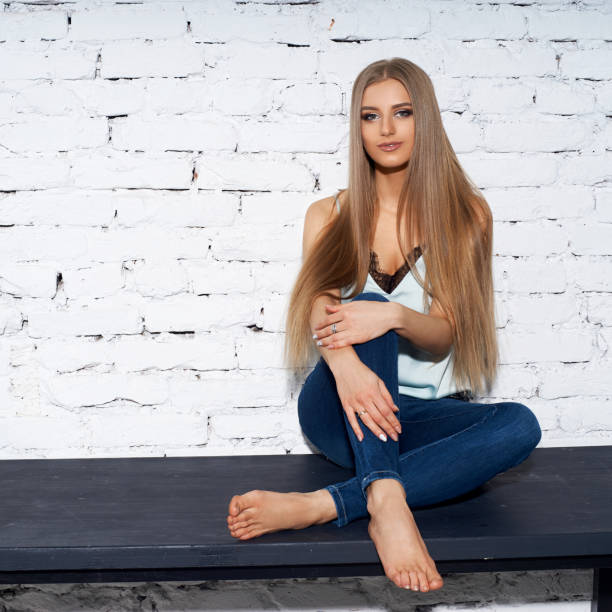Teenage girl in jeans, sneakers and black blouse is standing, holding hand on chin, looking away and smiling. Front view. Full length studio shot isolated on white.