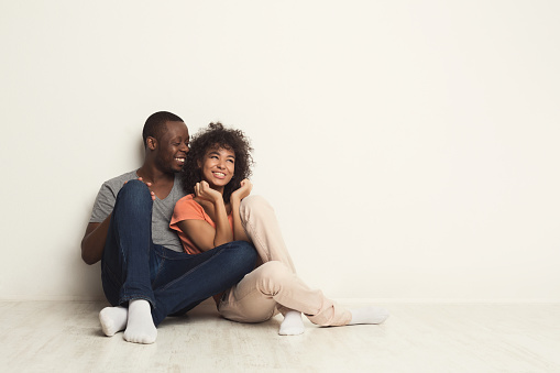 Happy african-american family couple hugging and talking, sitting on floor in new apartment, dreaming about future, copy space, isolated