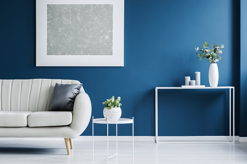 Metal console table with plant in vase and candles standing on blue wall in living room interior with light grey sofa and modern poster
