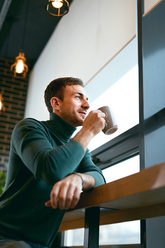 Man Drinking Coffee Drink In Cafe Near Window. Handsome Young Smiling Man In Stylish Clothes Holding Cup With Beverage In Hand In Cozy Cafe Indoors. High Quality Image