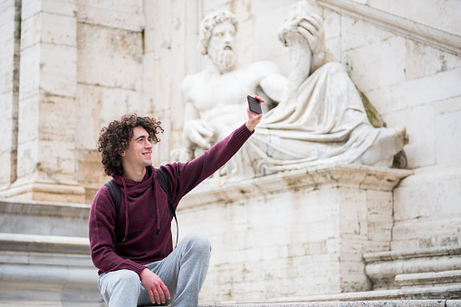 Handsome young man with curly hair in tracksuit taking selfie with his mobile phone in front of Nile God statue in Rome