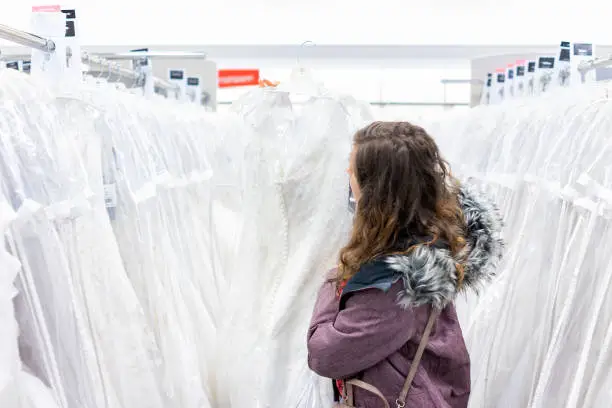 Young woman holding lace wedding dress in boutique discount store, many white garments hanging on rack hangers row