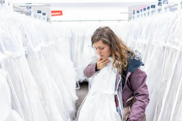 Young woman trying on lace wedding dress gown in boutique discount store, many white garments hanging on rack hangers row