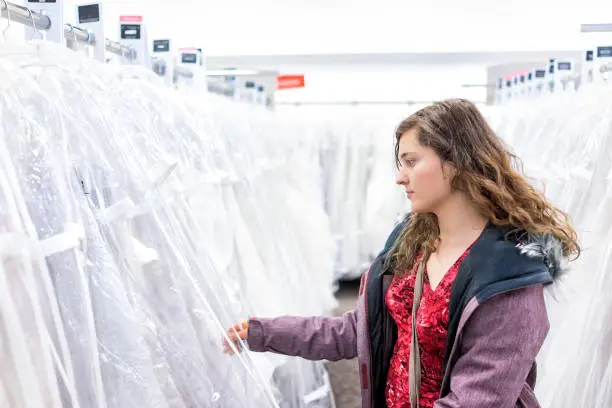 Young woman searching trying on touching wedding dress in boutique discount store, many white garments hanging on rack hangers row