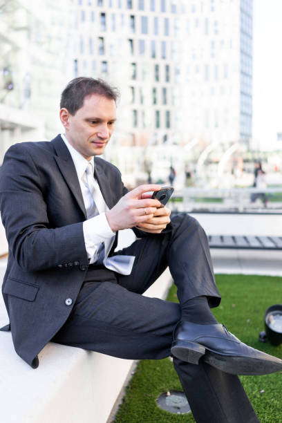 istock Young businessman sitting on bench in park, using smartphone phone mobile cellphone smiling looking down texting in suit and tie, cheerful on interview break 974832200