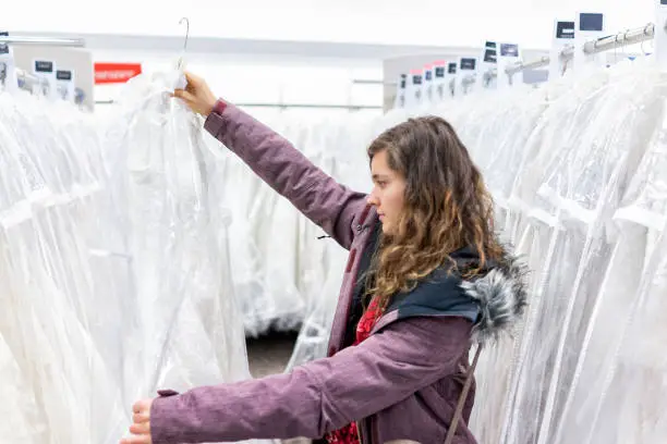 Happy young woman holding one lace wedding dress in boutique discount store, many white garments hanging on rack hangers row