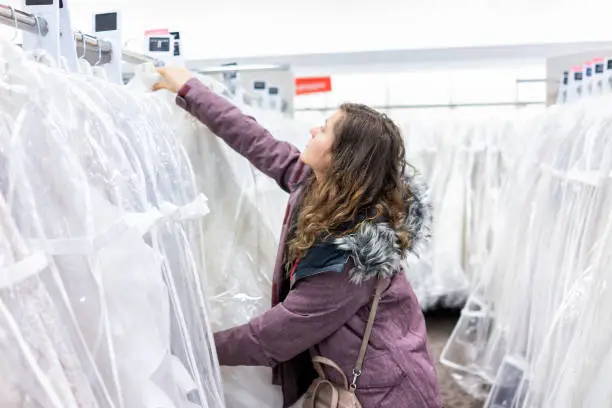 Young woman searching trying on touching one lace wedding dress in boutique discount store, many white garments hanging on rack hangers row