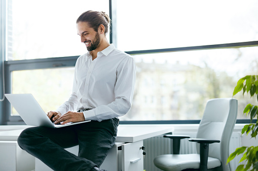 Man Working On Notebook In Office. Portrait Of Young Professional Smiling Businessman In In White Shirt With Laptop Working At Workplace In Modern Light Office. Business People. High Quality Image.