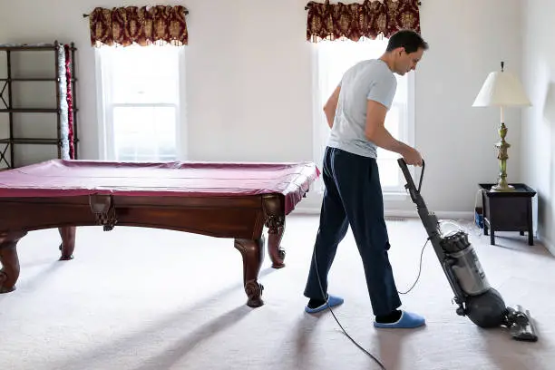 Young man house husband vacuuming using vacuum hoovering carpet floor inside interior of house living room billiards table, domestic life