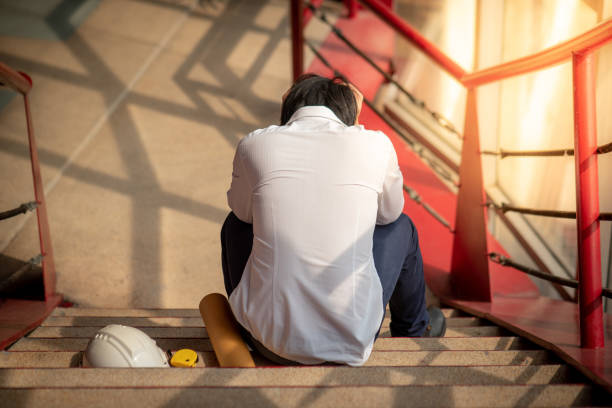 istock Engineer or Architect feeling stressed, worried and tired with his job while sitting on stair with protective safety helmet at construction site.  Problem and failure with construction project concept 974937252