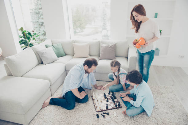 porträt von nachdenklichen vater und zwei kinder spielen schach brettspiel planungsstrategie sitzen auf teppich in der nähe von sofa, lächelnde mutter bringt teller mit obst für gamer. idyllische konzept - concentration chess playing playful stock-fotos und bilder