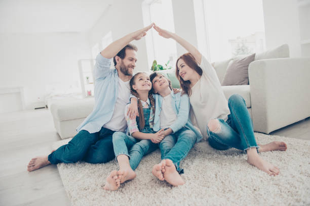 nouveau bâtiment résidentiel maison achat appartement concept. élégant complet famille avec deux enfants assis sur les tapis, papa et maman faisant figure de toit avec mains bras dessus de tête - sofa family fun child photos et images de collection