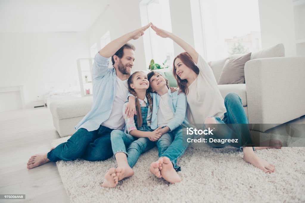 New building residential house purchase apartment concept. Stylish full family with two kids sitting on carpet, mom and dad making roof figure with hands arms over heads Family Stock Photo