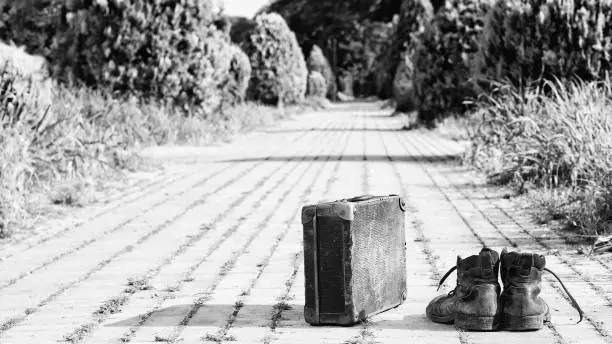 Old worn leather shoes and an antique cardboard suitcase with metal reinforcements at the corner, in the middle of a brick road. A black-and-white photo effect.