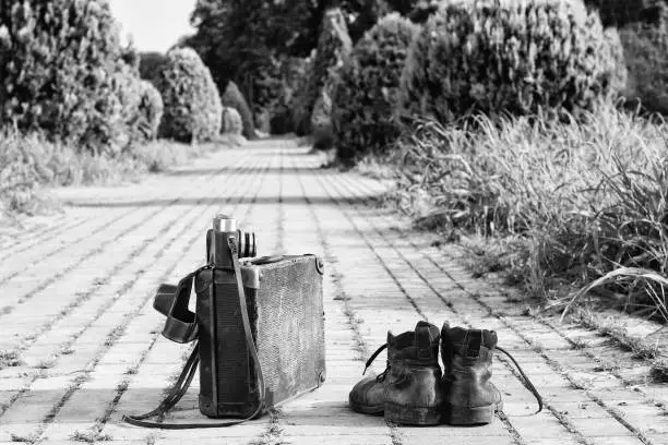 Traveling light! Worn ankle boots next to a vintage cardboard suitcase, a film camera in its open leather case, and a brick road. A black-and-white photo effect.