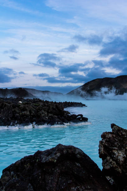 A view across a bright blue lagoon hot spring with black rocks at dusk in Iceland A wide angle image of a view across a hot spring in Iceland. The water in the spring is bright blue and the water is steaming around black rocks. The sun has nearly set and the sky is blue. blue lagoon iceland photos stock pictures, royalty-free photos & images