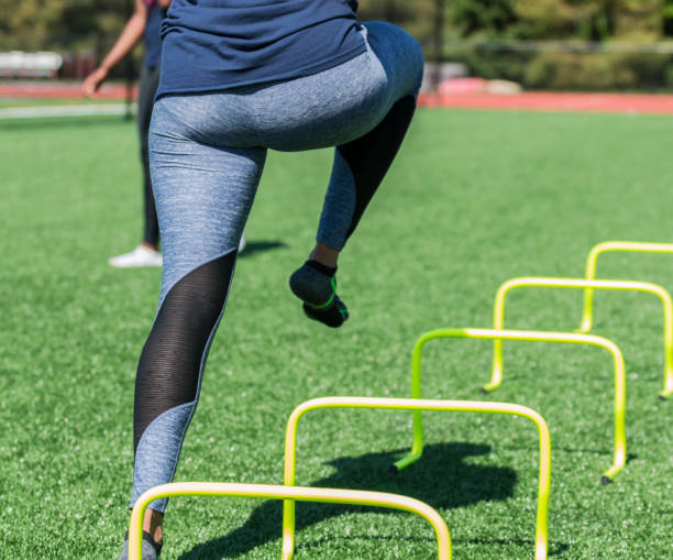 Running over yellow mini hurdles A female high school athlete performs running drills over yellow mini banana hurdles on a turf field with no shoes on, only socks in blue spandex. crossfit girl stock pictures, royalty-free photos & images