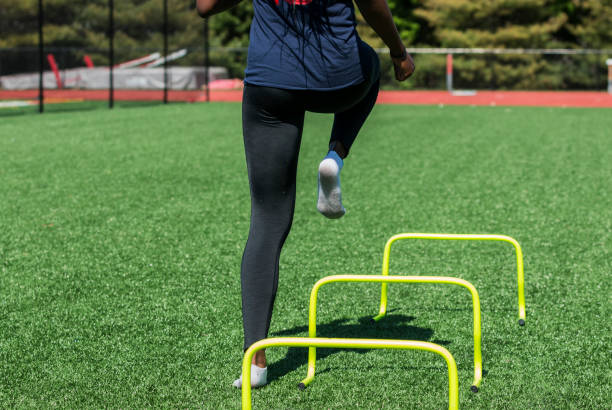 Female stepping over yellow hurdles A high school girl is performing speed and agility drills over hurdles with no shoes on, only socks on a green turf field wearing black spandex, viewed from behind. sports training drill stock pictures, royalty-free photos & images