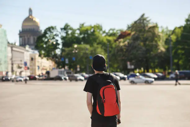 Back view of young stylish man tourist walking at Palace square near Saint Isaac's Cathedral in Saint-Petersburg. He wearing in black jeans, black T-shirt, black cap and red backpack.