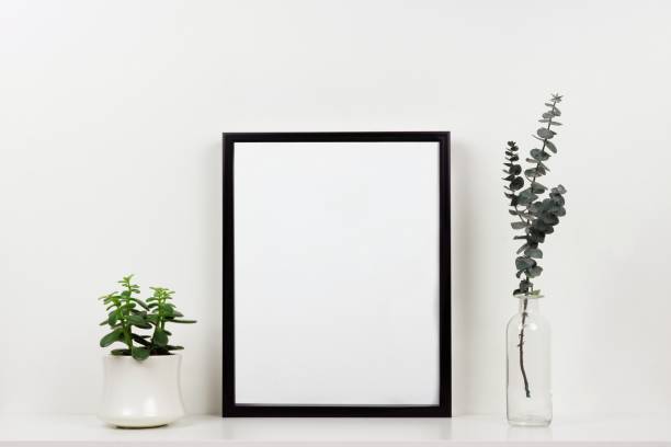 istock Mock up black frame against white wall with plant and branches on a white shelf 975274708
