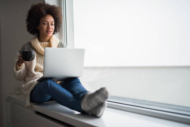 istock African American woman relaxing on window sill and using laptop. 975574818