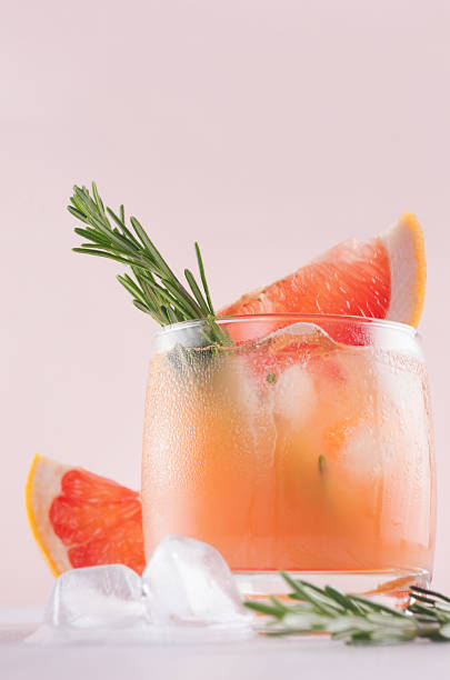 istock Refreshing cold citrus cocktail with ice cubes, green rosemary and slice grapefruit closeup on soft light pink and white background. 975637432