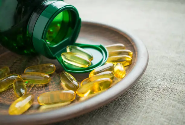 Photo of Yellow vitamin D3 (cholecalciferol) gelatine capsules and green bottle on clay plate on burlap background