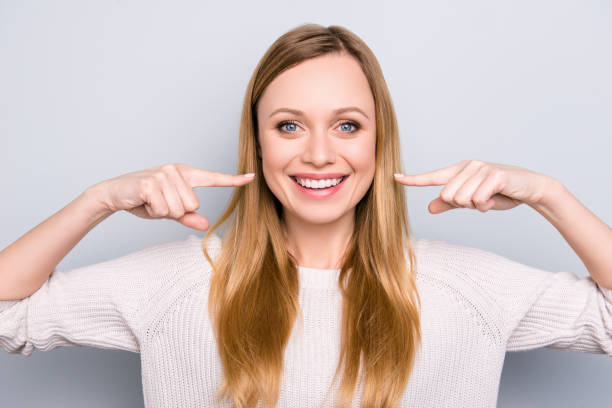 ritratto di gioiosa ragazza soddisfatta che gesticola i suoi traventi denti bianchi sani con due indice che guardano la fotocamera isolata su sfondo grigio. concetto ortodontico - sorriso aperto foto e immagini stock