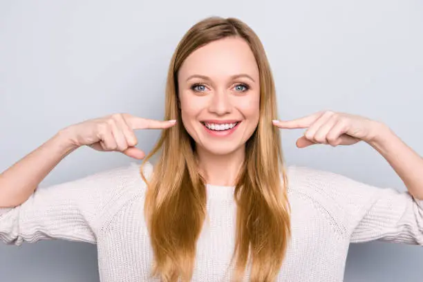 Photo of Portrait of joyful satisfied girl gesturing her beaming white healthy teeth with two forefingers looking at camera isolated on grey background. Orthodontic concept