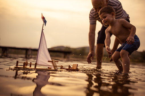 Grandfather and grandson playing with a sailboat toy on the lake