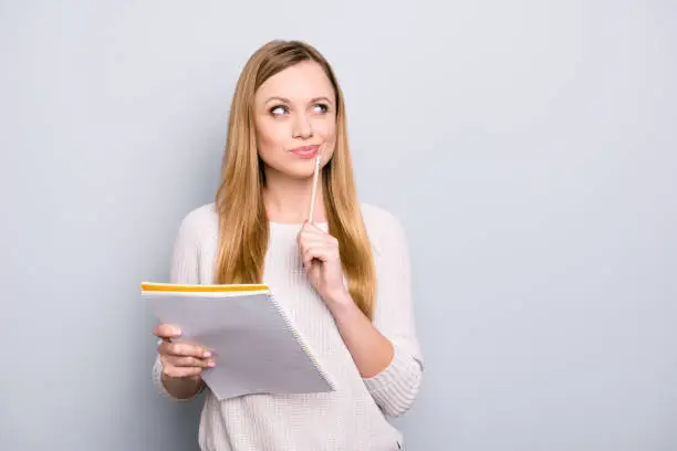 Photo of Portrait with copy space empty place of thoughtful pensive girl having copybook and pen in hands looking up, writer waiting for muse, isolated on grey background