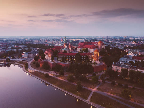 Krakow, Poland - May 14, 2018: Illuminated royal Wawel castle and cathedral at night and light reflections in Vistula River. Riverside with park, trees, promenade, lanterns, harbor and tourist ship