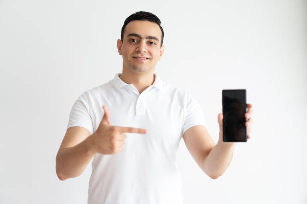 istock Smiling young man pointing at smart phone and looking at camera 975705460