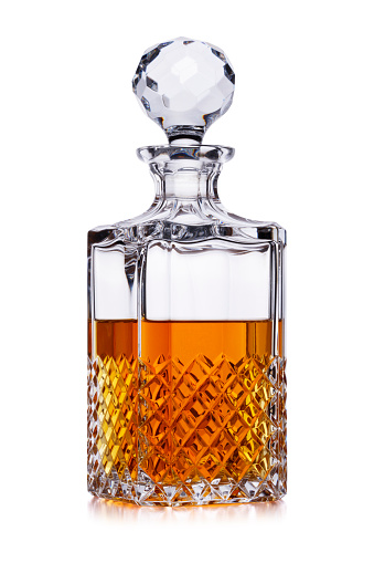 A half full crystal whisky decanter, with amber alcohol, shot on white, with a small reflection