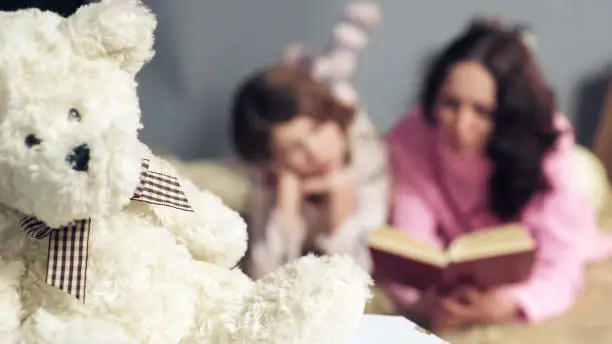 Photo of Closeup of fluffy toy, mom and daughter reading fairy tales on background