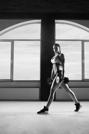 Black and white photo athletic woman keeping dumbbells in big, spacy hall. Having strong, fit body with heatlthy tanned skin and muscles. Doing fitness exercises. Wearing modern sportswear.