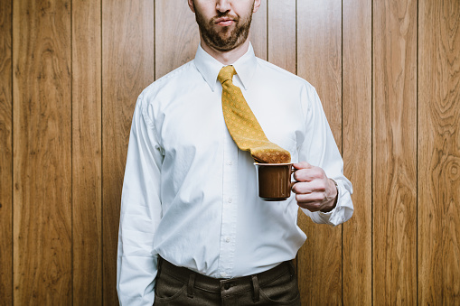 A business man in an office environment looks disappointed that his necktie has fallen in to his cup of coffee.  Workplace problems and humor.