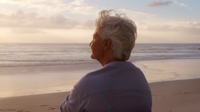 Rear View Of Senior Woman On Beach Watching Sun Set Over Ocean