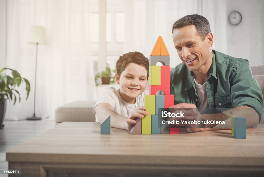 Glad father and son constructing house from small details Happy man and boy are playing together at home. They are building toy tower and laughing. Friendly family concept Father Stock Photo