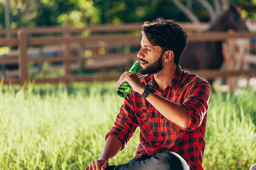 Young farmer sitting on bench and drinking beer with pasture and corral in background