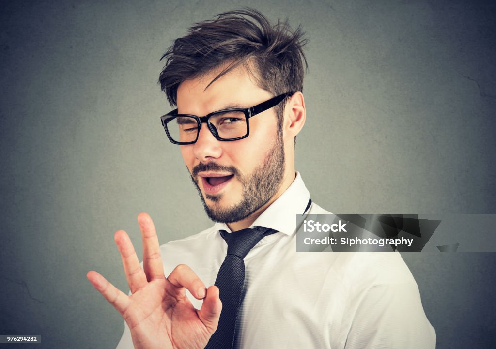 Ironic man showing OK gesture Young bearded man in glasses showing OK gesture and blinking at camera with overconfidence Humor Stock Photo