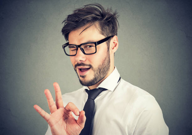 Ironic man showing OK gesture Young bearded man in glasses showing OK gesture and blinking at camera with overconfidence young man wink stock pictures, royalty-free photos & images