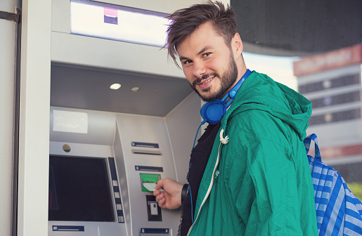 Young hipster guy in headphones smiling at camera while inserting credit card in ATM machine getting cash money