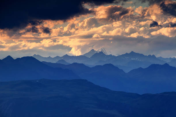 Jagged blue ridge silhouettes, snowcapped Rotspitze Pizzo Rosso peak and fluffy orange clouds at sunset, Venediger Group and Villgraten Mountains Defereggen Alps Hohe Tauern Osttirol Austria Europe