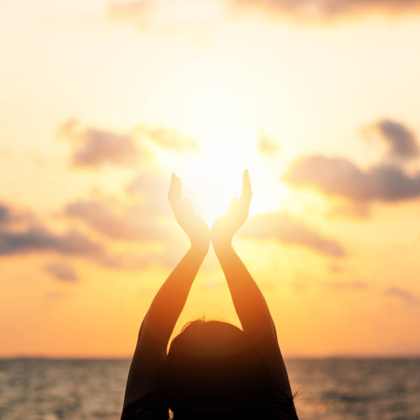 summer sun solstice concept et la silhouette des mains de jeune femme heureuse se détendre, de méditer et de tenant du coucher du soleil dans le ciel l’heure d’or chaud sur la plage avec le fond de l’océan ou la mer - day sunlight sky sea photos et images de collection