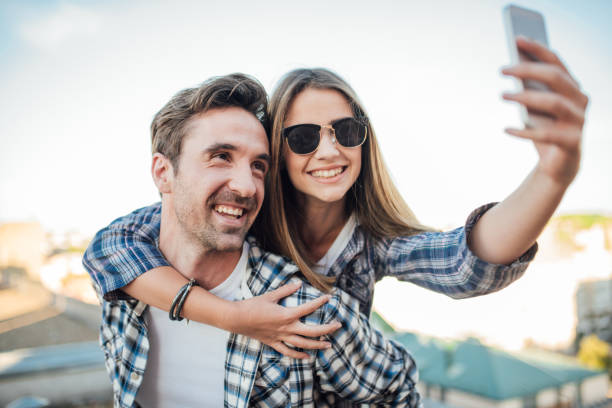 istock Man holding woman with sunglasses on his back while she is taking a selfie 976563658