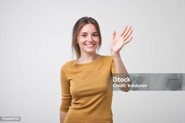 Friendlylooking European Teenager Dressed In Yellow Pulover Saying Hello Waving Her Hand Stock Photo - Download Image Now