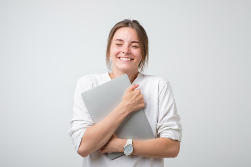 Cheerful caucasian woman in white t-shirt holding laptop computer and smiling. She is glad to complete a project or job