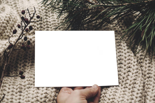 istock christmas card mockup. hand holding empty christmas card with space for text, template on knitted sweater and fir branches.  merry christmas, seasonal greetings, happy holidays. 976610802
