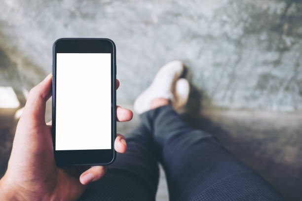 istock Mockup image of hand holding black mobile phone with blank white screen  with white canvas shoes on concrete polishing wall 976637916